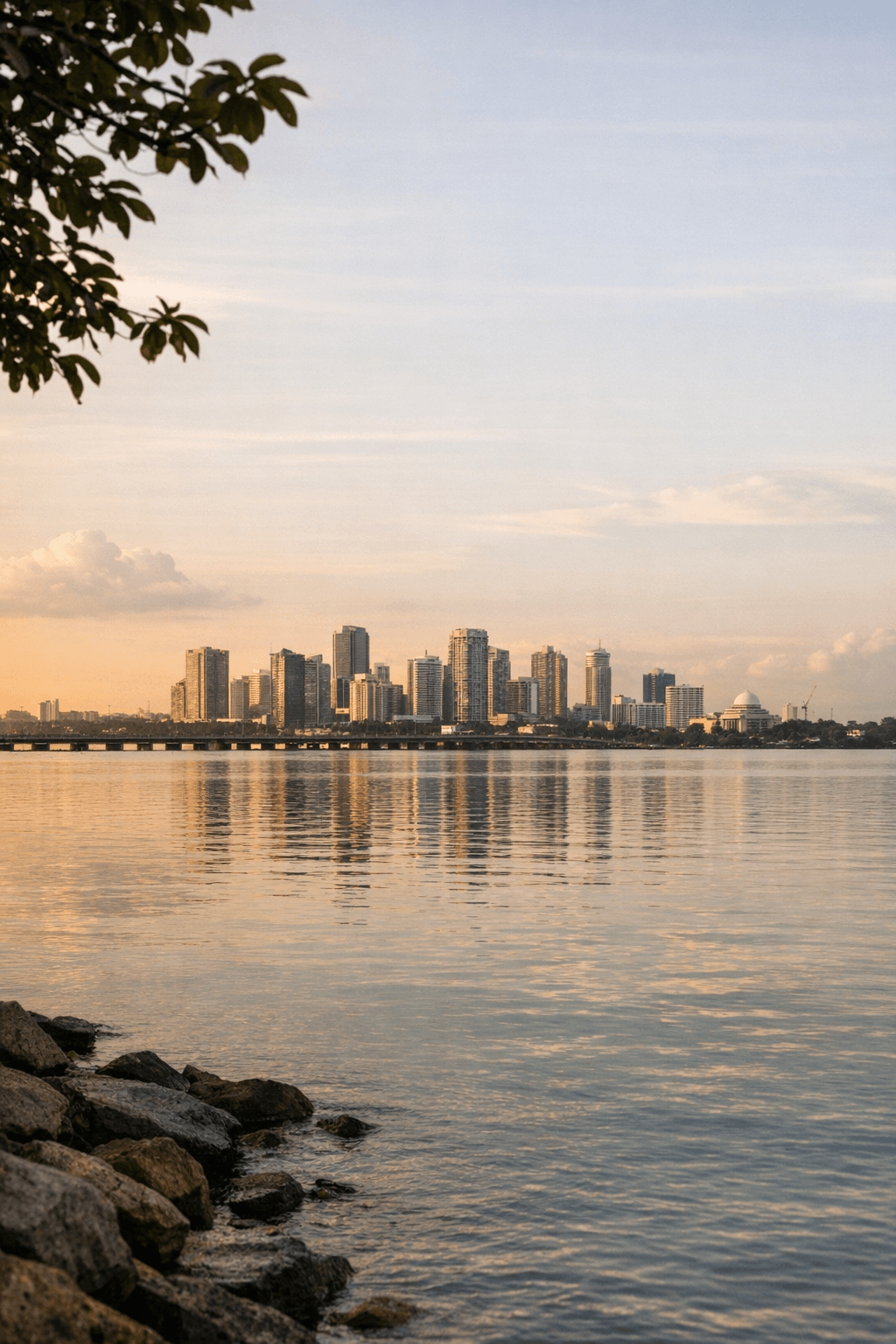 Golden-hour view across the Straits of Johor from the Singapore side toward the distant Johor Bahru skyline, the Causeway a faint line across calm water