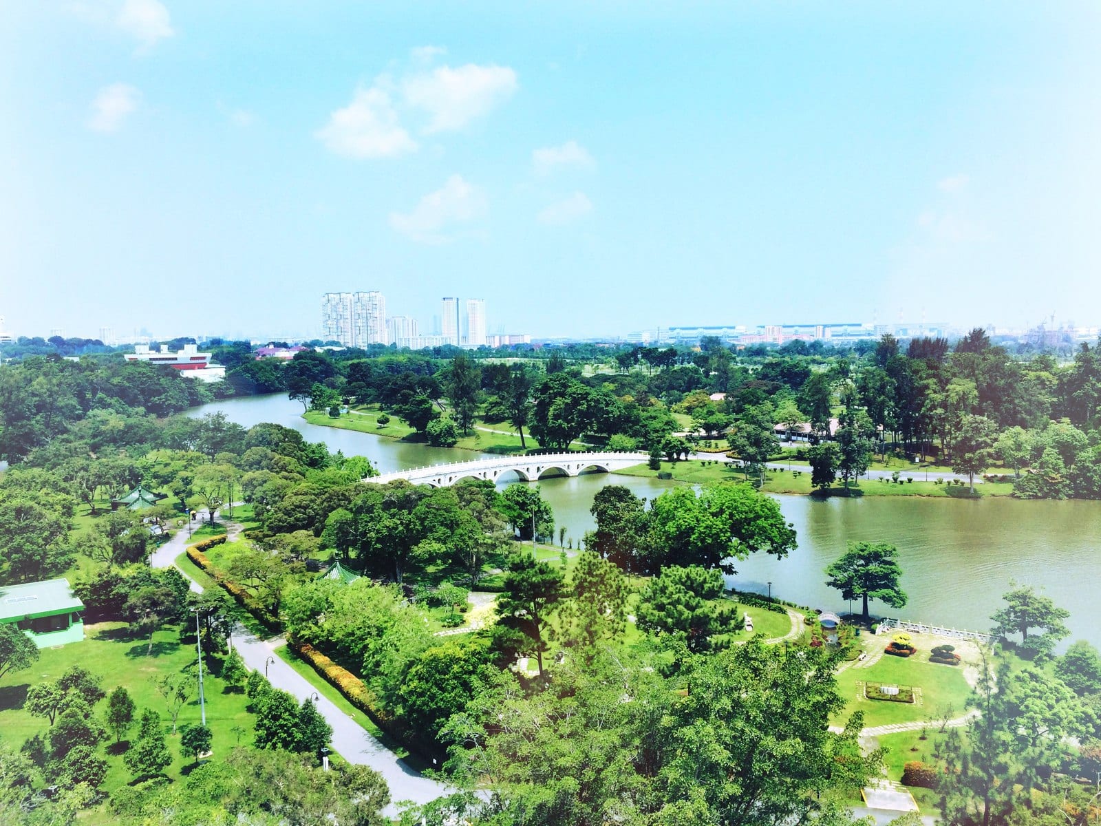Aerial view of Jurong Lake Gardens — the lake, the white arched bridge to the Chinese Garden and lush parkland — with the Jurong skyline beyond, the green setting beside Sora in District 22.